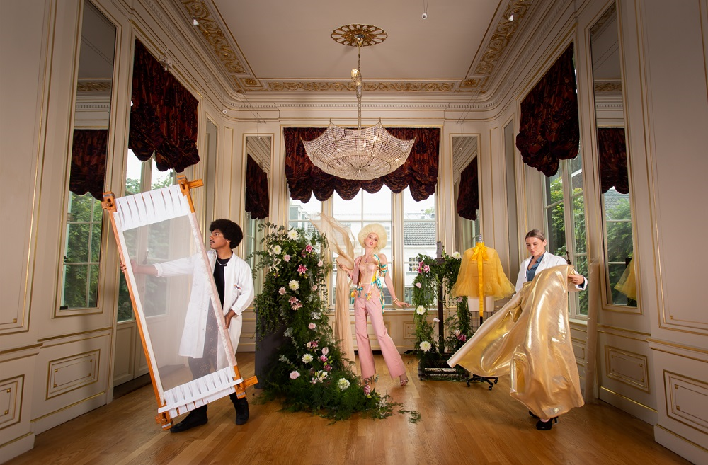 Scène in paleiszaal van opbouw tentoonstelling met kroonluchter, bloemdecoraties en model in zachtroze kleding